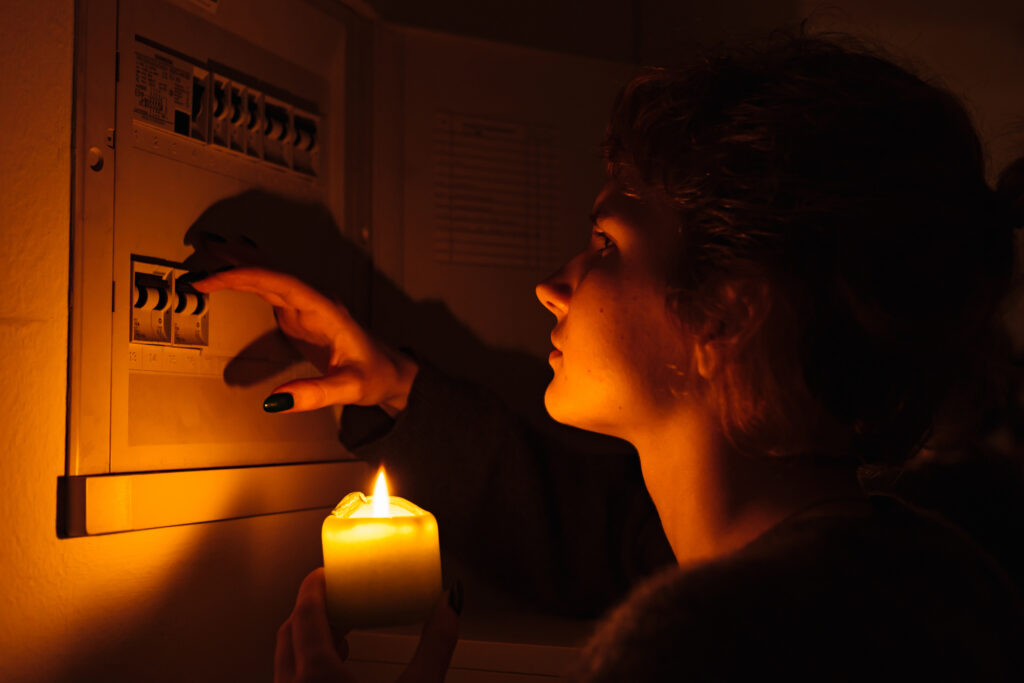 In complete darkness, a woman examines the fuse box or electrical distribution board at home during a power outage, relying on candles for light. With blackout conditions and no electricity, she urgently needs lightning.